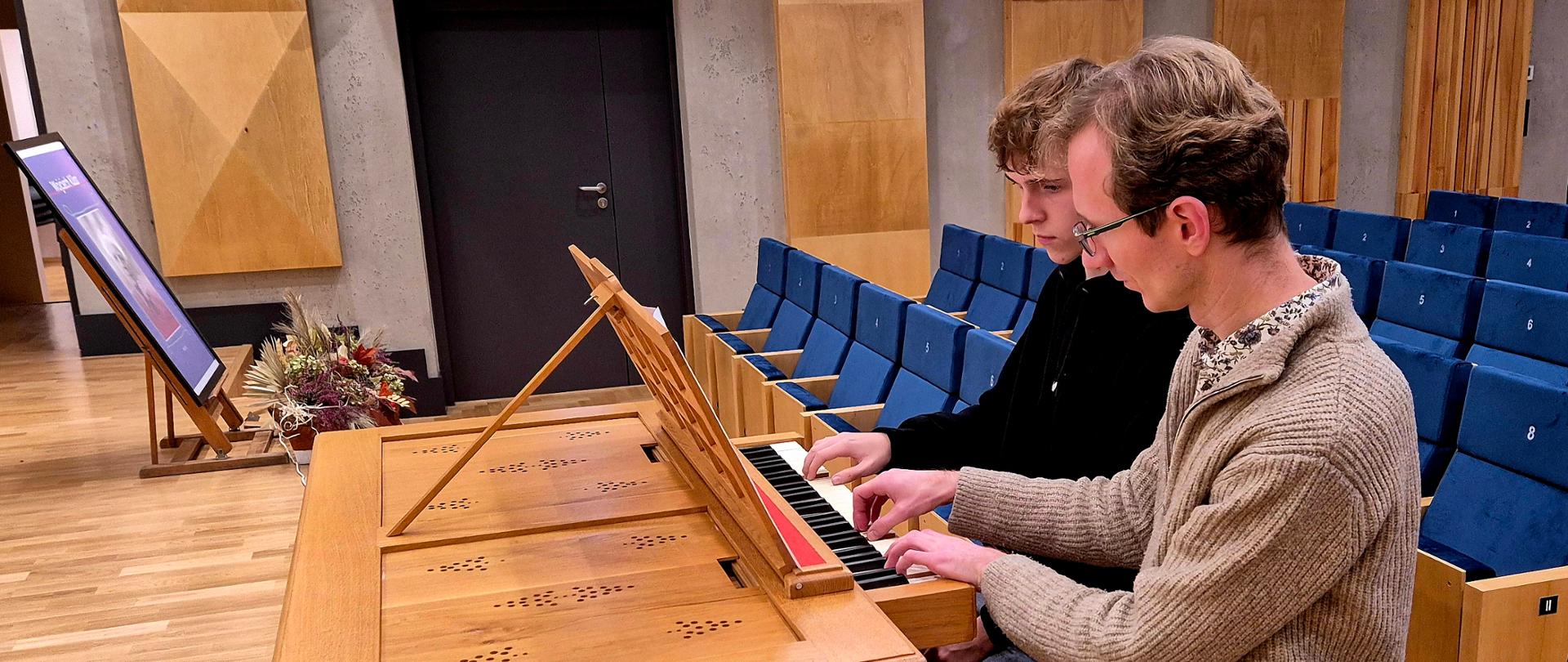 Zdjęcie przedstawia nauczyciela i ucznia siedzących przy pozytywie na sali koncertowej