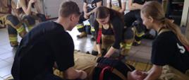 Zdjęcie przedstawia kursantów podczas udzielania pierwszej pomocy. Ratownicy ubrany w ubranie specjalne i rękawiczki jednorazowe. Obok znajdują się uczestnicy kursu.