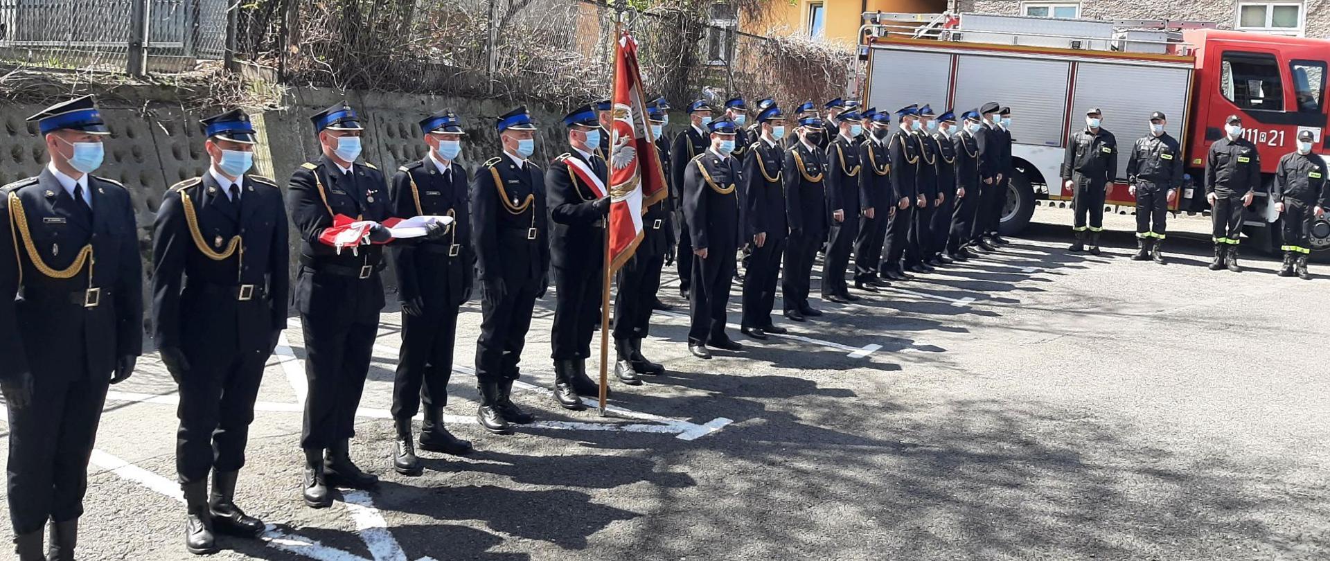 Kolorowa fotografia wykonana na zewnątrz, obok zabudowań, w pogodny dzień. Przedstawia na pierwszym planie pododdział umundurowanych na galowo, ze sznurami galowymi i czapkami rogatywkami na głowach, strażaków na zbiórce w dwuszeregu, z pocztem flagowym trzymającym złożoną flagę biało-czerwoną, pocztem ze sztandarem jednostki. Zbiórka na placu asfaltowym w maseczkach na twarzach. W tle czerwony pojazd bojowy straży pożarnej, drzewa i widoczna zabudowa miejska.