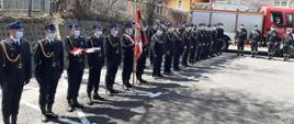 Kolorowa fotografia wykonana na zewnątrz, obok zabudowań, w pogodny dzień. Przedstawia na pierwszym planie pododdział umundurowanych na galowo, ze sznurami galowymi i czapkami rogatywkami na głowach, strażaków na zbiórce w dwuszeregu, z pocztem flagowym trzymającym złożoną flagę biało-czerwoną, pocztem ze sztandarem jednostki. Zbiórka na placu asfaltowym w maseczkach na twarzach. W tle czerwony pojazd bojowy straży pożarnej, drzewa i widoczna zabudowa miejska.