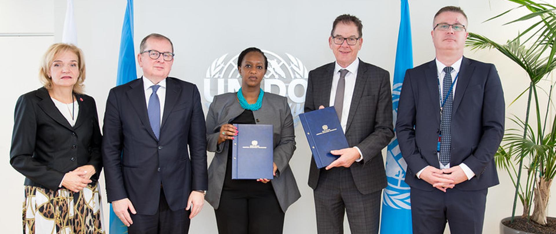 Pięć osób stoi razem w pomieszczeniu przed tłem z logo UNIDO (Organizacji Narodów Zjednoczonych ds. Rozwoju Przemysłowego). Dwie osoby w środku trzymają niebieskie teczki. Po bokach stoją dwie flagi ONZ i Ukrainy. Wszyscy są formalnie ubrani i patrzą w stronę aparatu.