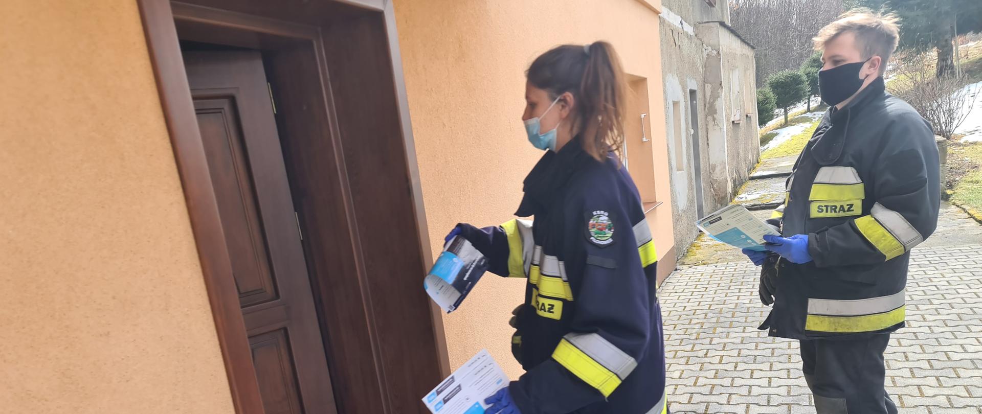 Zdjęcie przedstawia dwóch strażaków z jednostki Ochotniczej Straży Pożarnej w Pilchowicach, którzy roznoszą ulotki informacyjne dotyczące szczepień przeciwko Covid-19. Strażacy znajdują się przed budynkiem mieszkalnym. Zdjęcie obrazuje moment przekazania ulotek mieszkańcom.
