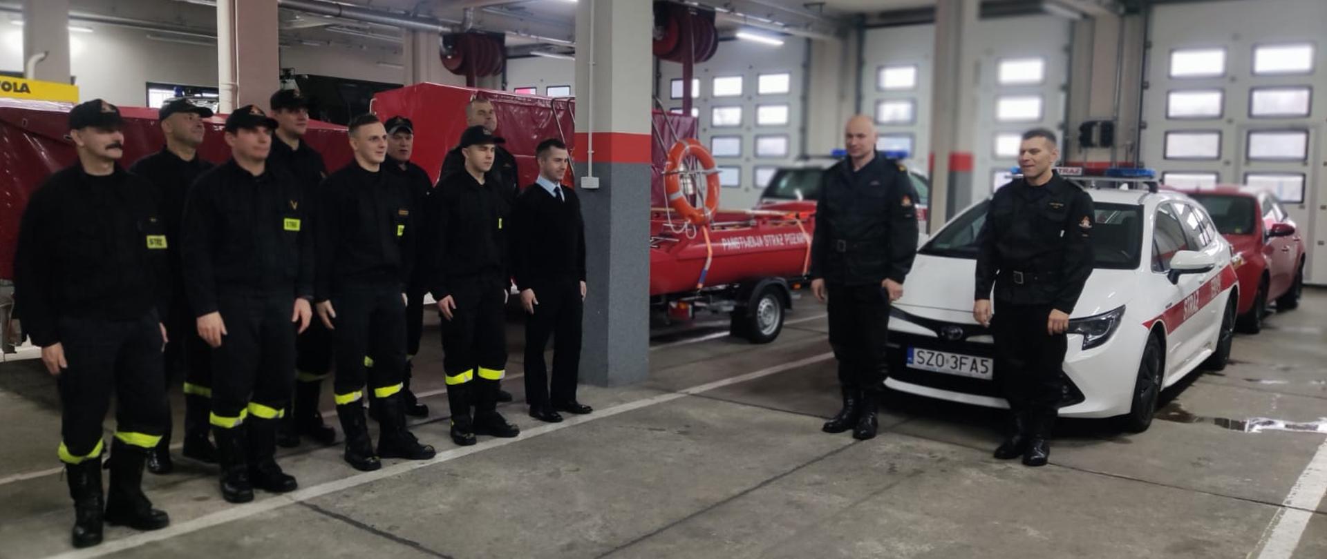 Po lewej stronie zdjęcia strażacy stojący w dwuszeregu na garażu. Za nimi samochody pożarnicze oraz inny sprzęt strażacki. Na środku dwóch strażaków stoi poza szeregiem.