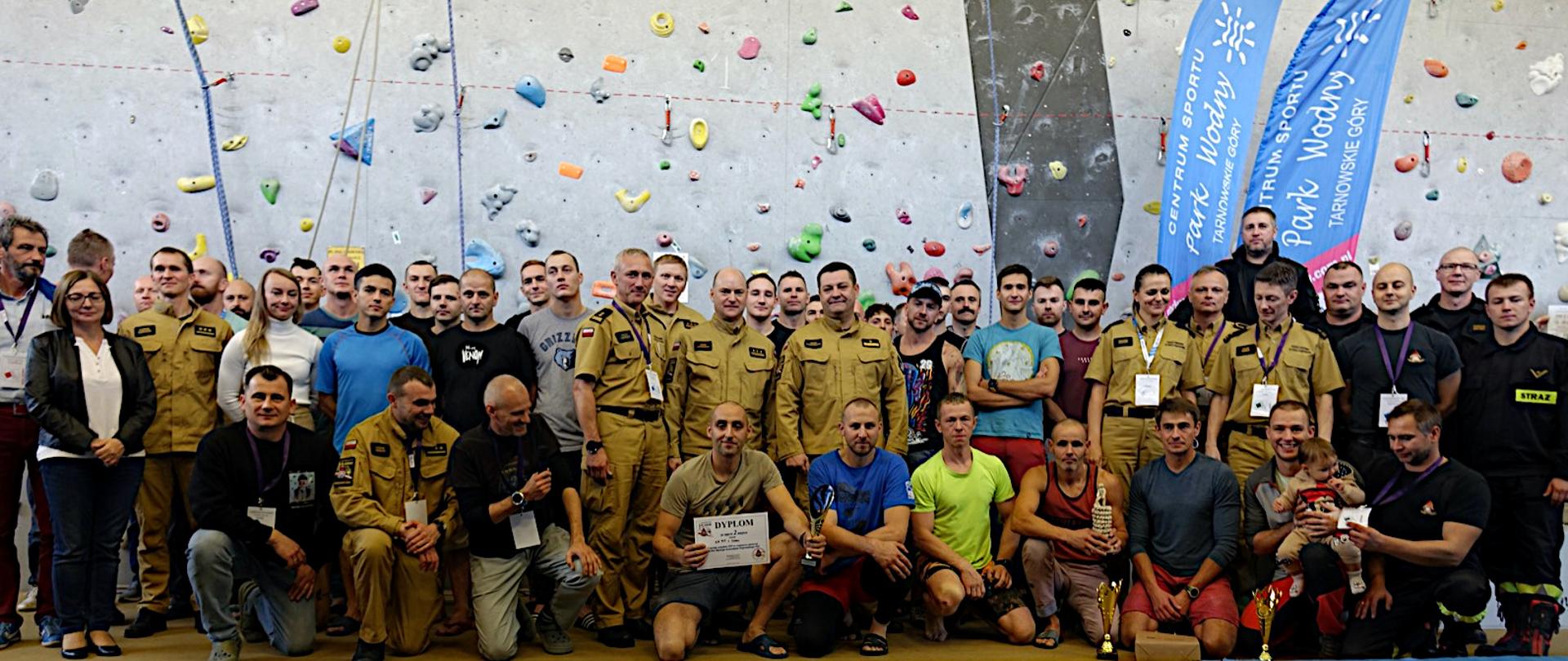 Grupa zawodników w strojach sportowych i strażaków w mundurach służbowych i ubraniach koszrowych ustawiona na tle ścianki wspinaczkowej, pozuje do zdjęcia grupowego.