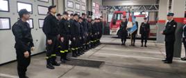 W lewej części zdjęcia strażacy stojący w dwuszeregu, w jego centralnej części poczet sztandarowy a za nim drabina pożarnicza, w prawej części zdjęcia przemawiający Komendant. 