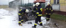 Pożar smarta. Parking przy Orliku w Bojanowie. Na pierwszym planie spalony pojazd i strażacy. W tle ogrodzenie posesji i budynek. Wokół samochodu pozostałości piany gaśniczej. 