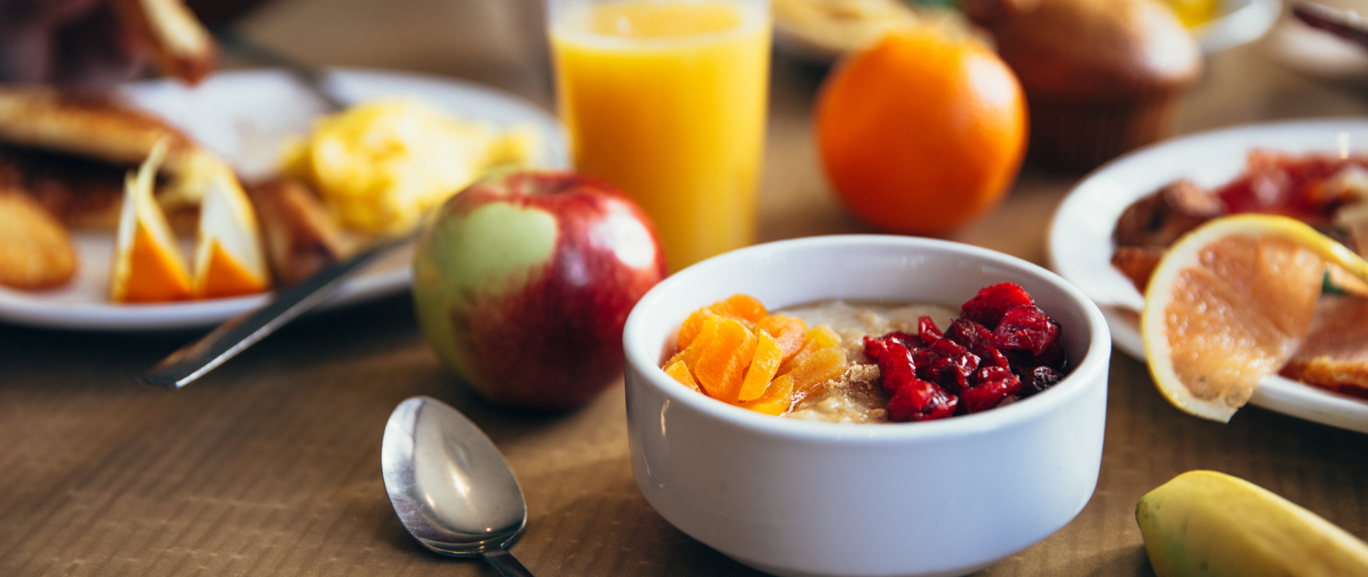 Na zdjęciu widać miskę z płatkami i owocami. Obok miski leży łyżka. W tle widać owoce