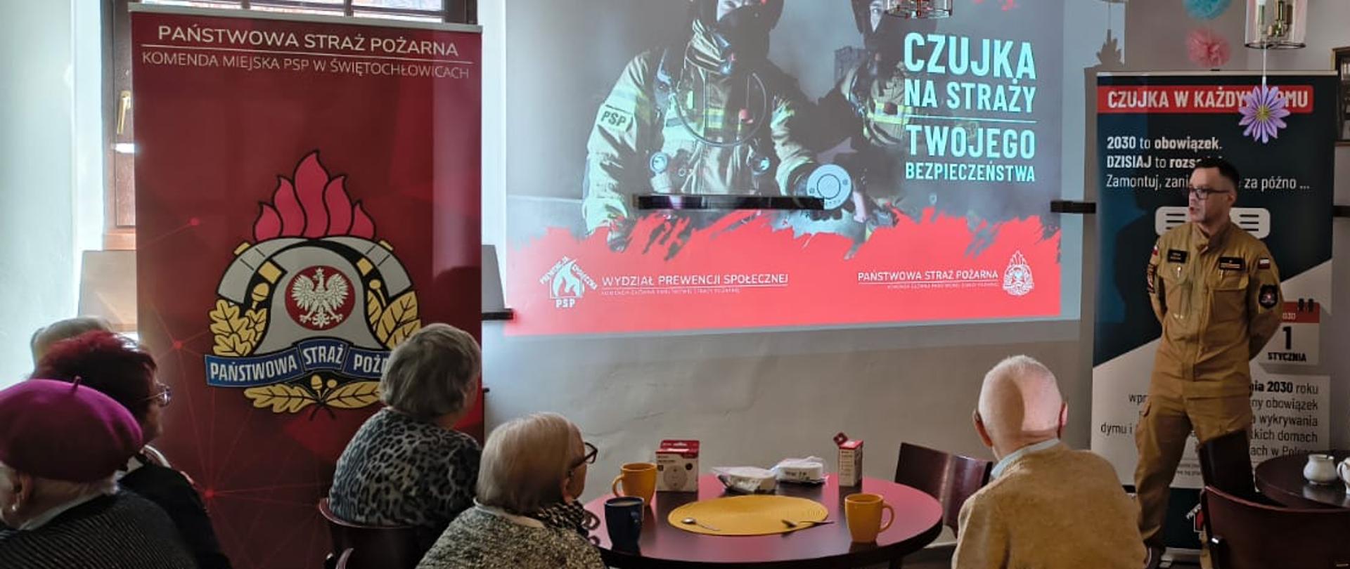 Na zdjęciu uwieczniono spotkanie edukacyjne prowadzone przez funkcjonariuszy Państwowej Straży Pożarnej w Świętochłowicach. Jeden strażak w piaskowym mundurze służbowym prezentuje materiały szkoleniowe grupie seniorów siedzących przy okrągłych stolikach. W tle widoczny jest ekran z prezentacją dotyczącą sygnałów alarmowych oraz baner informacyjny kampanii „Czujka na straży Twojego Bezpieczeństwa”, co sugeruje, że tematyka spotkania dotyczy szeroko pojętego bezpieczeństwa przeciwpożarowego i obrony cywilnej.