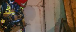 Zdjęcie przedstawia strażaka w pomieszczeniu wędzarni podczas sprawdzania ukrytych zarzewi ognia przy pomocy kamery termowizyjnej. Strażak ubrany w pisakowe ubranie specjalne, czerwony hełm wraz z założonym aparatem oddechowy.