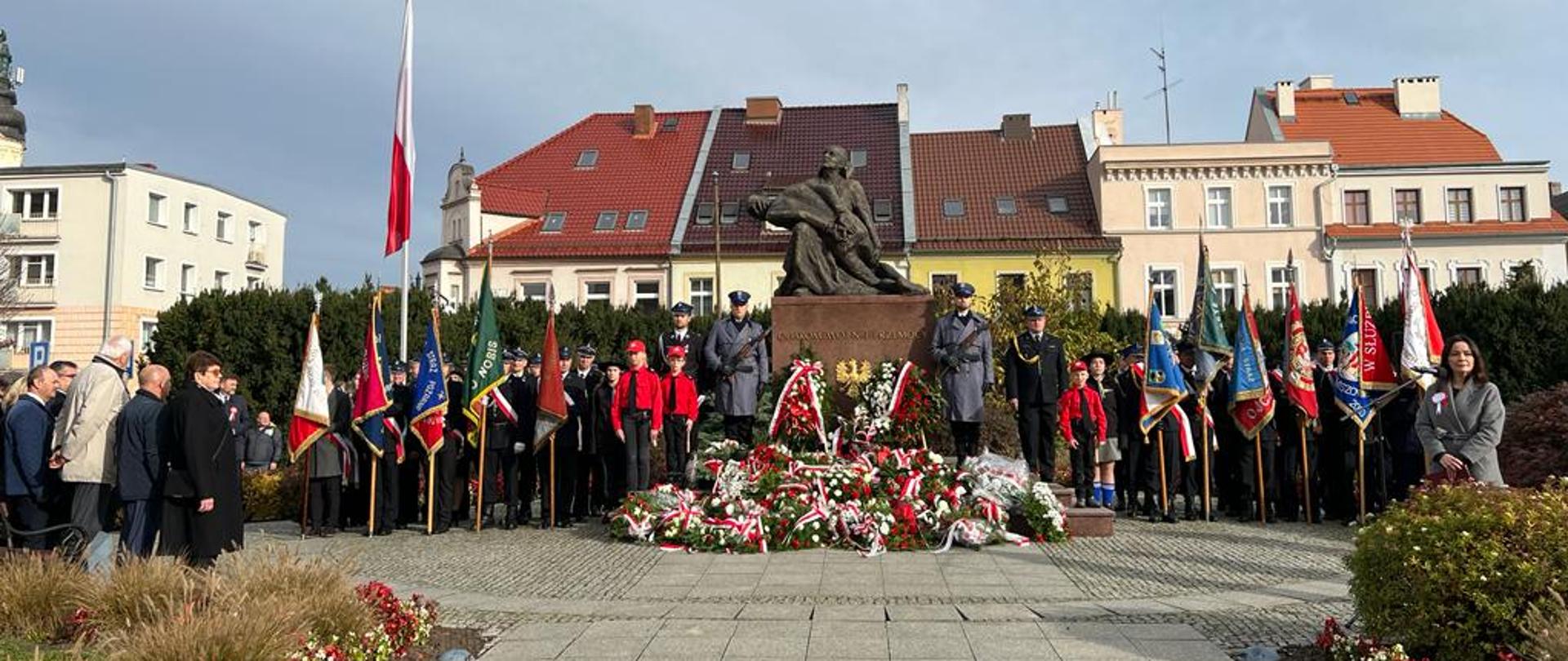 Na zdjęciu znajdują się osoby ubrane w mundury, poczty flagowe ustawione przy pomniku, w tle widać budynki