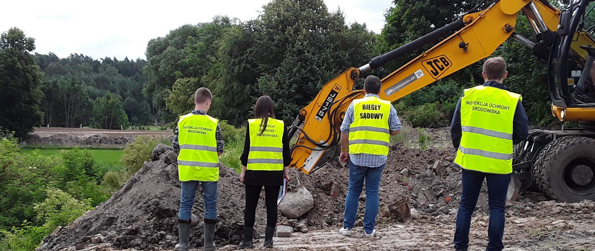 Inspektorzy z Biegłym Sądowym w żółtych kamizelkach stoją przy pracującej koparce, która wykopuje odpady. W tle zielony las.