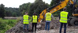 Inspektorzy z Biegłym Sądowym w żółtych kamizelkach stoją przy pracującej koparce, która wykopuje odpady. W tle zielony las.