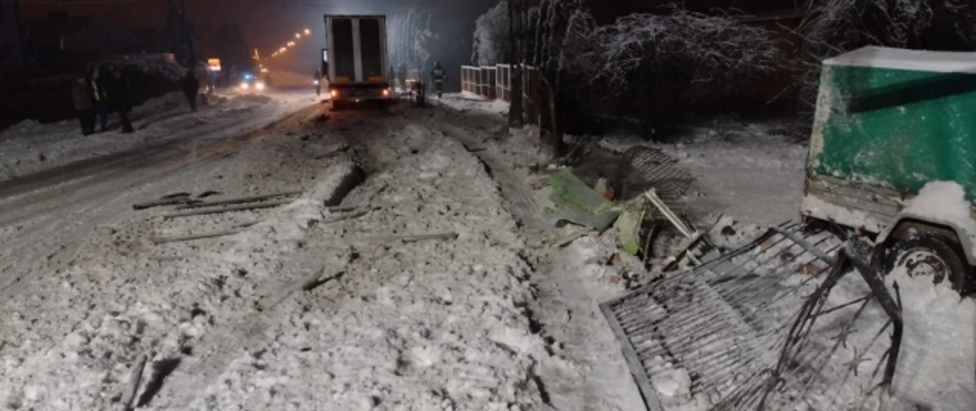  Na pierwszym planie uszkodzone metalowe przęsła ogrodzenia. W oddali naczepa samochodu ciężarowego. W tle nocna zimowa aura.