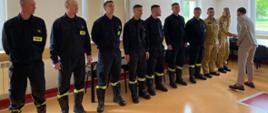 Zdjęcie przedstawia grupę strażaków ustawionych w szeregu wewnątrz jasnego pomieszczenia szkoleniowego. Większość z nich ubrana jest w czarne ubranie koszarowe z widocznym napisem „STRAŻ”, a dwóch strażaków po prawej stronie nosi beżowe mundury specjalne. Po prawej stronie kadru Prezydent Ciechanowa w beżowym garniturze wita się z jednym ze strażaków. W tle znajdują się okna, stoły oraz zawieszony pod sufitem projektor multimedialny