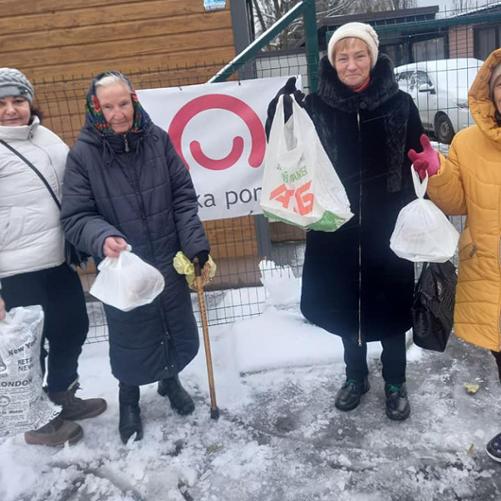 Grupa kobiet przed budynkiem pozuje do zdjęcia, trzymając w rękach torby z jedzeniem