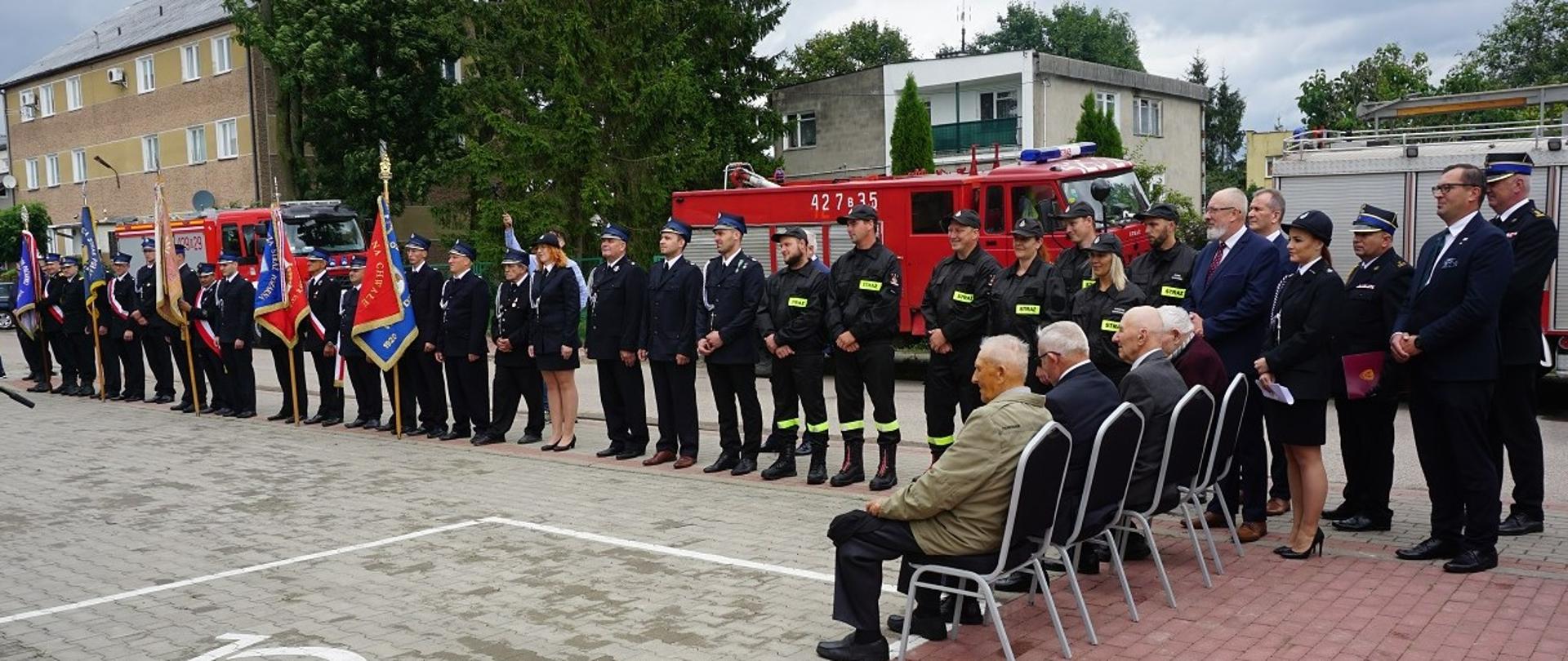Zaproszeni goście stoją przed remizą OSP. W tle samochód strażacki.