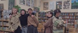 We wnętrzu biblioteki (regały, prace malarskie i rysunkowe na ścianach) stoi pięć dziewcząt trzy po lewej, dwie po prawej obrócone do siebie plecami 