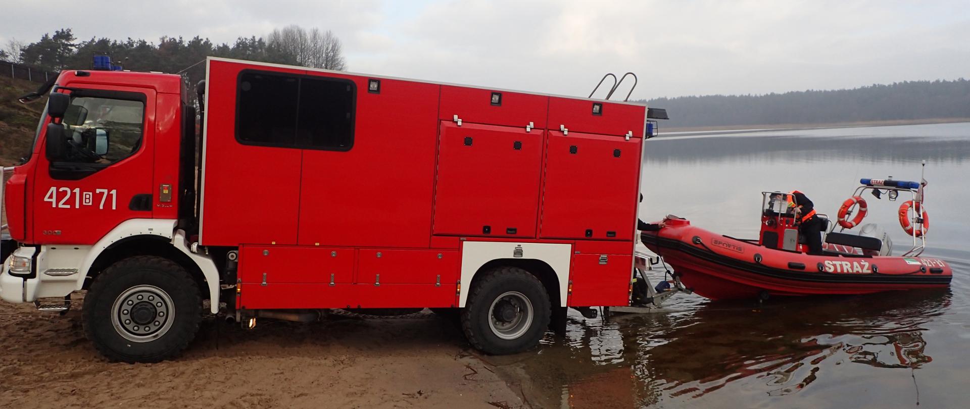 Na zdjęciu samochód ratownictwa wodnego Renault Midlum 300 koloru czerwonego woduje łódź strumieniową hybrydową Sportis z piaszczystego brzegu jeziora. Na łodzi strażak w czarnym ubraniu koszarowym z założoną kamizelką asekuracyjna w kolorze pomarańczowym. W tle jezioro oraz las.