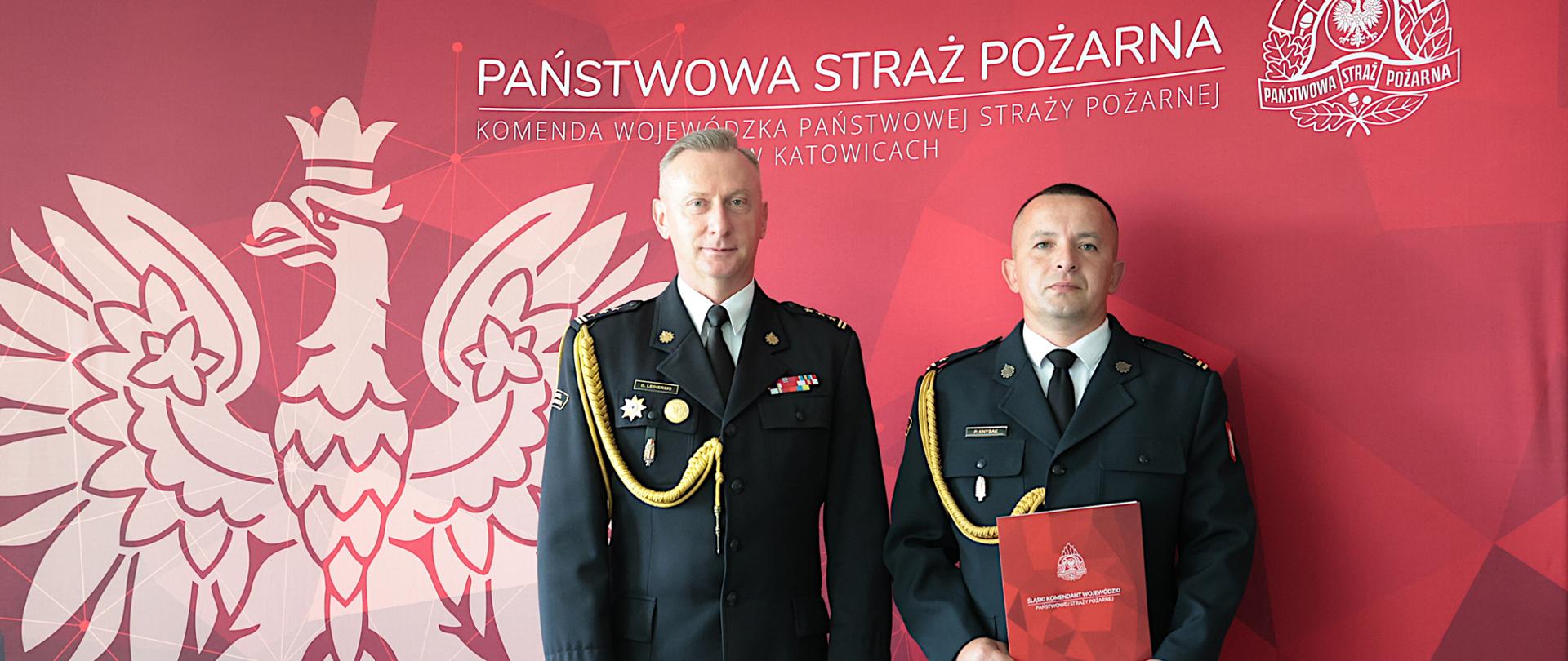 Na zdjęciu widać dwóch strażaków w mundurach wyjściowych Państwowej Straży Pożarnej stojących obok siebie na tle czerwonej ścianki z dużym białym wizerunkiem orła oraz napisem „Państwowa Straż Pożarna – Komenda Wojewódzka Państwowej Straży Pożarnej w Katowicach”. W prawym górnym rogu znajduje się emblemat PSP.
Po lewej stronie stoi śląski komendant wojewódzki PSP - st. bryg. Damian Legierski Po prawej powołany na stanowisko zastępcy komendanta miejskiego PSP w Częstochowie – mł. bryg. Piotr Knysak, który trzyma przed sobą czerwoną teczkę z godłem PSP oraz napisem „Śląski Komendant Wojewódzki Państwowej Straży Pożarnej”.