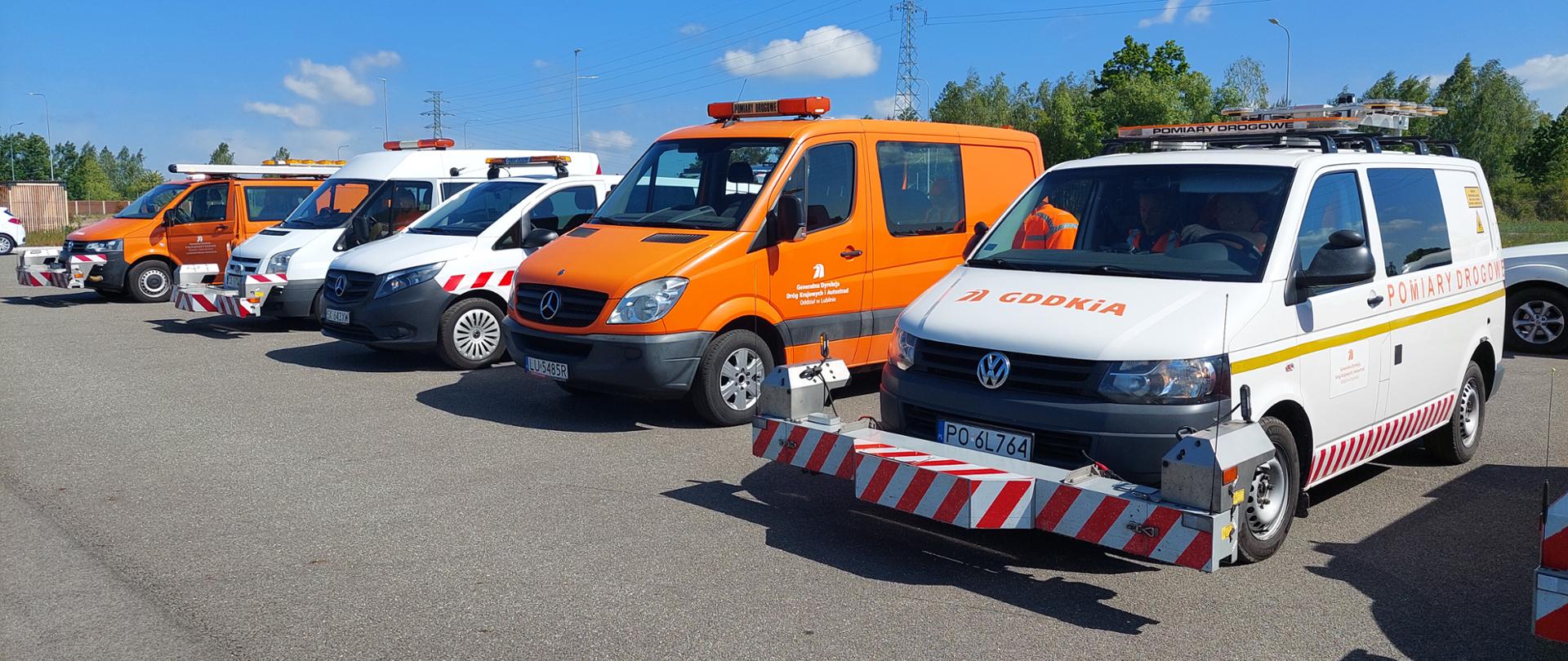 Kilka pojazdów technicznych, w tym białe i pomarańczowe vany z zamontowanymi z przodu urządzeniami pomiarowymi oznaczonymi czerwono‑białymi pasami, ustawionych w szeregu na otwartym placu przy słonecznej pogodzie.