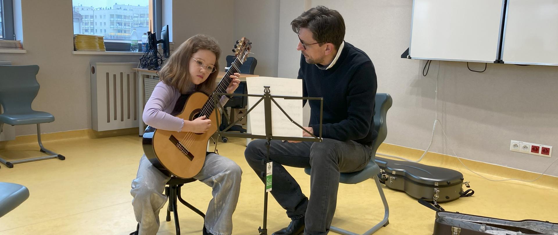 I Festiwal Gitarowy Lublin w dn. 2 marca 2024. Trwa lekcja gitary z uczennicą naszej szkoły Amelią Lewkowicz, którą prowadzi Tomasz Zawierucha profesor z Niemiec. Oboje siedzą na krzesłach, Amelia po lewej stronie gra na gitarze, przed nimi stoi pulpit z nutami. Wybija się na zdjęciu żółta podłoga.
