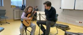 I Festiwal Gitarowy Lublin w dn. 2 marca 2024. Trwa lekcja gitary z uczennicą naszej szkoły Amelią Lewkowicz, którą prowadzi Tomasz Zawierucha profesor z Niemiec. Oboje siedzą na krzesłach, Amelia po lewej stronie gra na gitarze, przed nimi stoi pulpit z nutami. Wybija się na zdjęciu żółta podłoga.
