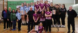 Grupowe zdjęcie uczennic i opiekunów po zawodach sportowych w sali gimnastycznej; drużyny stoją na podium i trzymają dyplomy