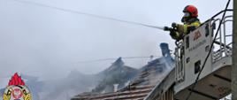 Zdjęcie przedstawia strażaka gaszącego palący się dach. Strażak znajduje się w koszu drabiny strażackiej i gasi wodą palące się drewniane elementy dachu.
