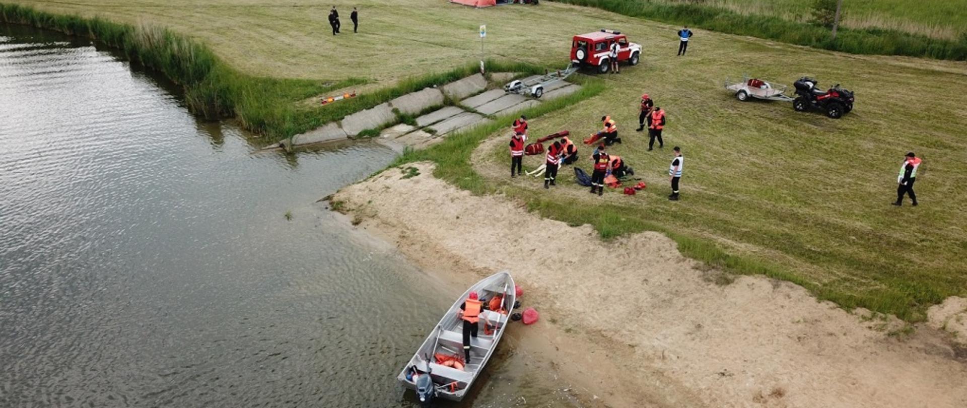 Na zdjęciu grupa strażaków udzielająca na brzegu zbiornika kwalifikowanej pierwszej pomocy osobom poszkodowanym wyciągniętym z wody