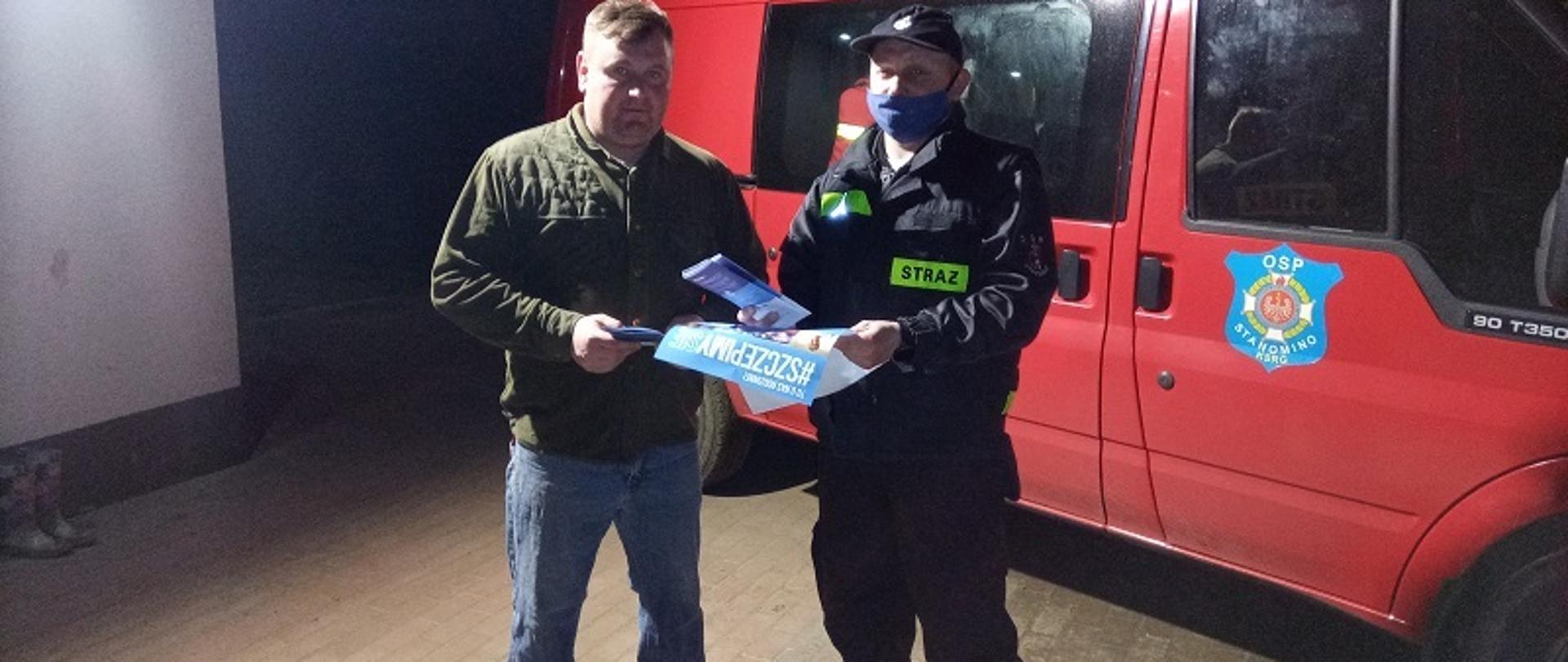Zdjęcie przedstawia dwie osoby stojące na tle samochodu strażackiego. Z lewej strony stoi osoba cywilna, która otrzymuje od druha OSP w ubraniu koszarowym ulotki i materiały informacyjne akcji #SZCZEPIMYSIĘ