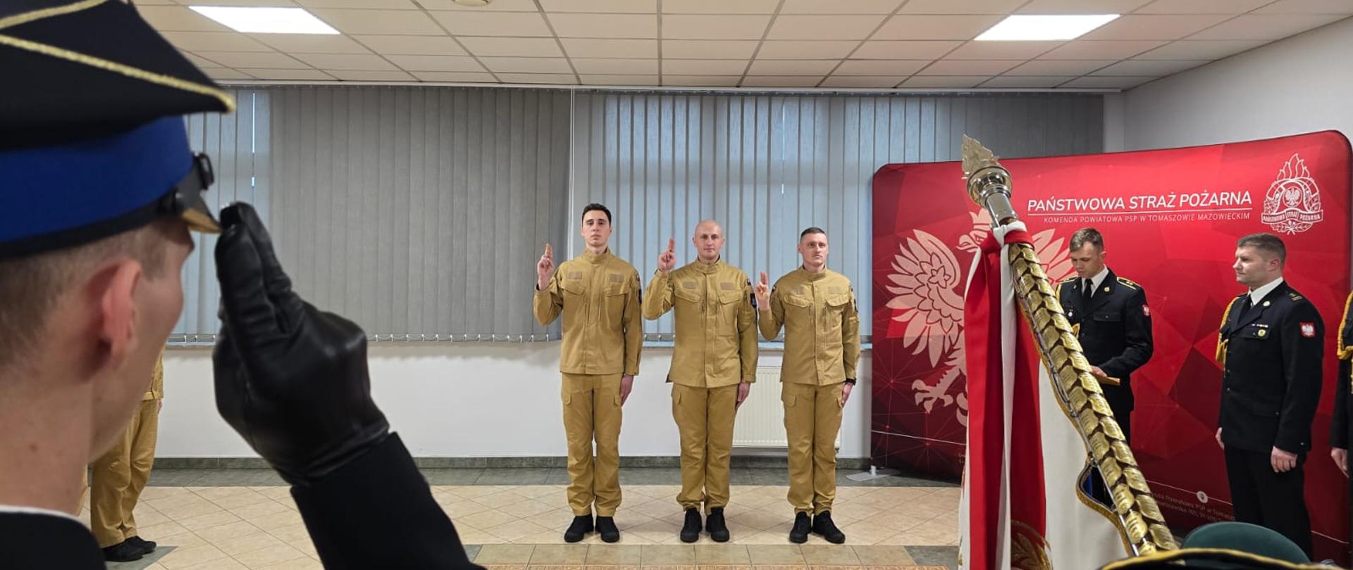 Trzech strażaków w ubraniach koloru piaskowego z wyciągniętymi dwoma palcami na wysokości ramion składa ślubowanie. Na pierwszym planie widać drzewiec sztandaru oraz strażaka w mundurze i czapce rogatywce który salutuje. 