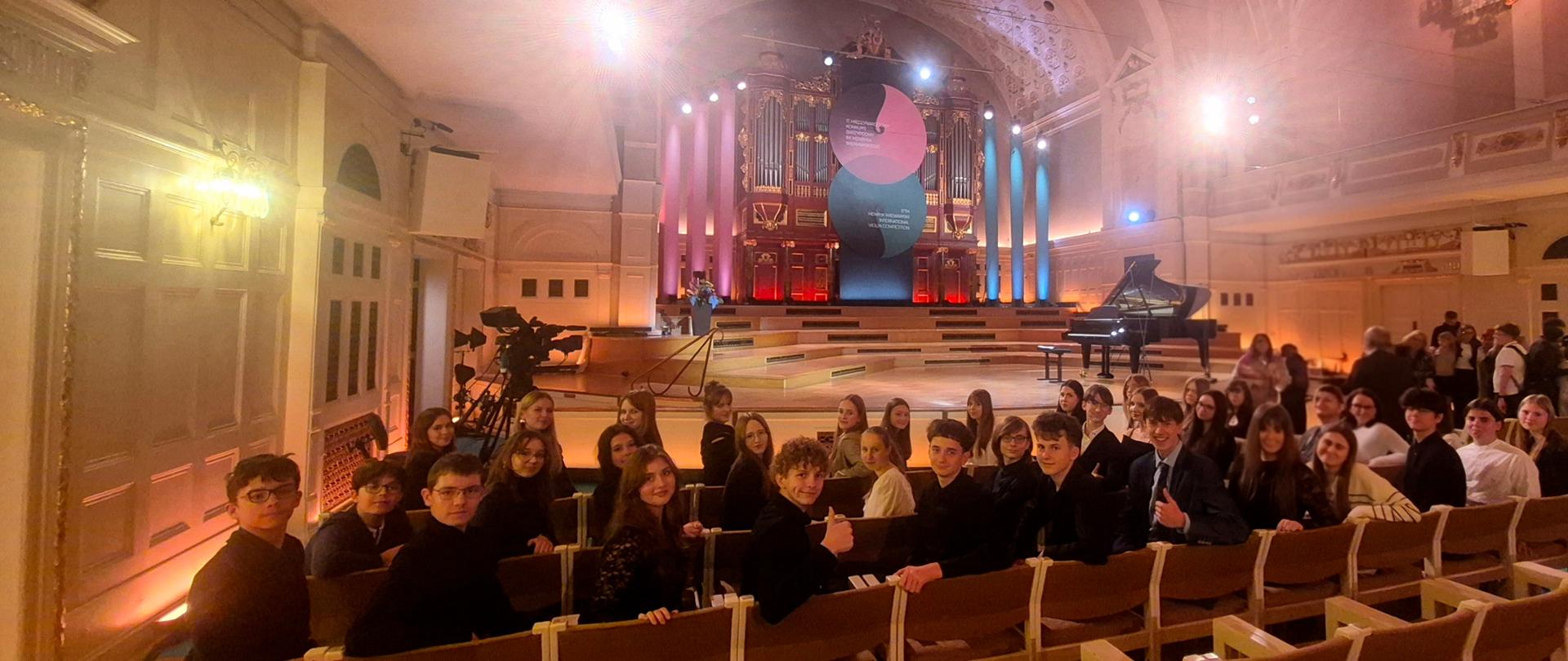 Grupa uczniów i nauczycieli siedzi na krzesłach odwrócona w stronę fotografa. W tle scena z fortepianem i dekoracje konkursowe