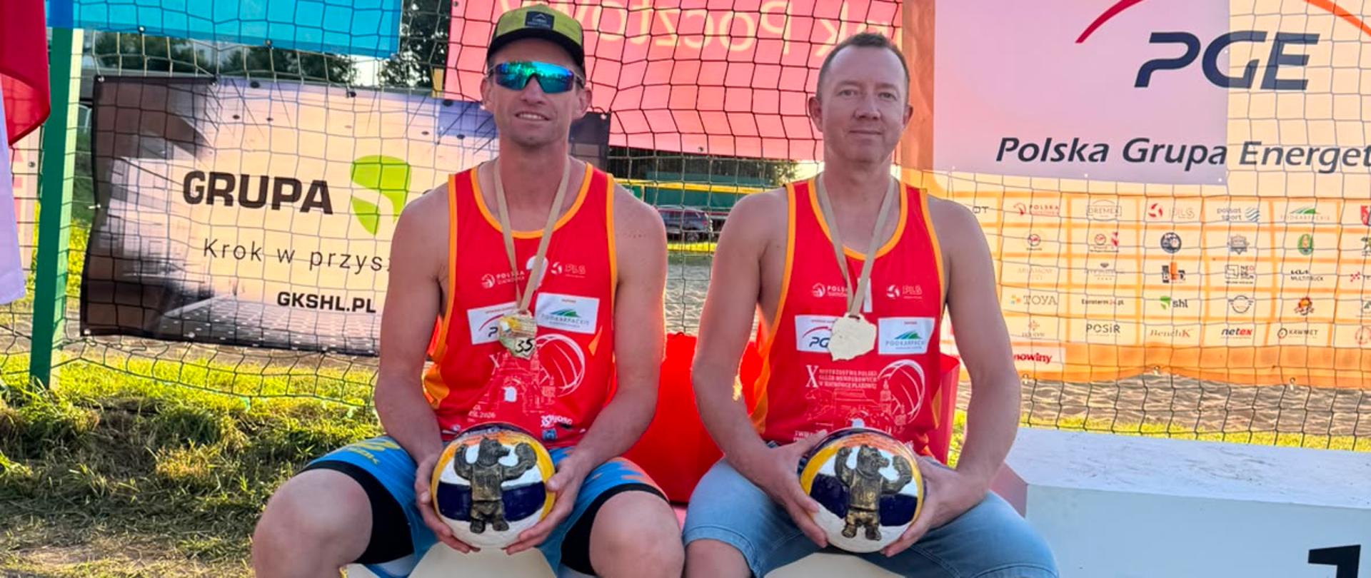 Dwaj zawodnicy siedzą na podium na piaszczystym boisku. Mają na sobie czerwone koszulki bez rękawów, krótkie spodenki i klapki. Każdy trzyma piłkę do siatkówki i ma na szyi złoty medal. Za nimi znajduje się siatka oraz duży baner z nazwą turnieju: X Mistrzostwa Polski Służb Mundurowych w Siatkówce Plażowej Twierdza Przemyśl 2026. W tle widoczne są logotypy sponsorów.