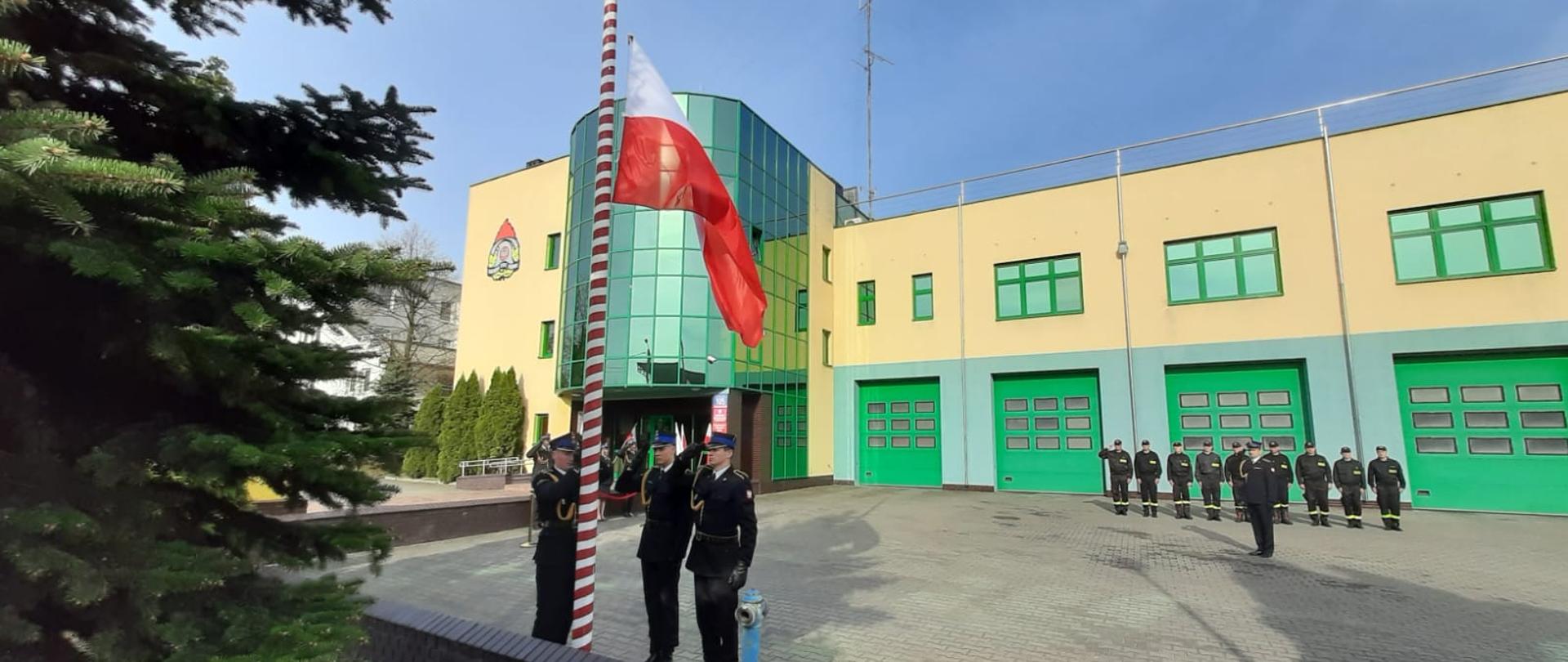 Trzech strażaków stoi przy biało czerwonym maszcie. Na maszcie powiewa biało czerwona flaga. W tle budynek strażnicy. Przed budynkiem ustawieni jeden obok drugiego stoją strażacy. 