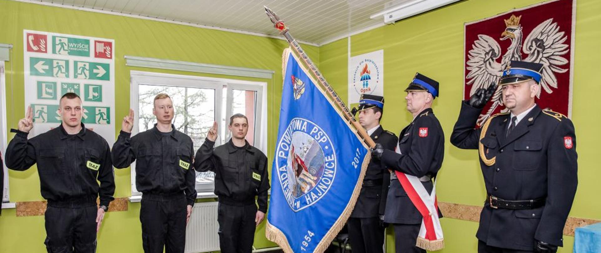 W pomieszczeniu z zielonymi ścianami stoją strażacy w czarnych mundurach. Jeden z nich trzyma sztandar. Dwaj salutują a trzech trzyma podniesione dwa palce w geście do ślubowania.