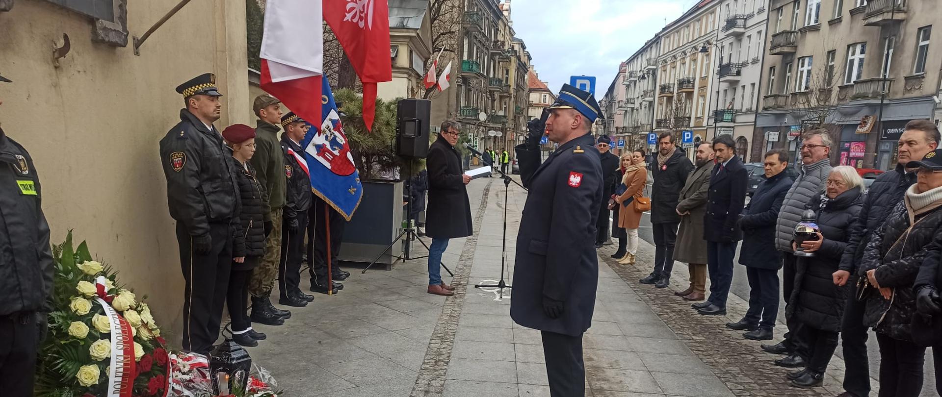Na zdjęciu widzimy strażaka ubranego w płaszcz galowy koloru granatowego, rogatywka na głowie. Strażak salutuje do tablicy pamiątkowej przymocowanej do ściany. Pod tablicą znajdują się kwiaty i wieniec w biało czerwonych barwach. W tle widzimy osoby przyglądające się wydarzeniom. Dalej widzimy ulice oraz budynki mieszkalne. 
