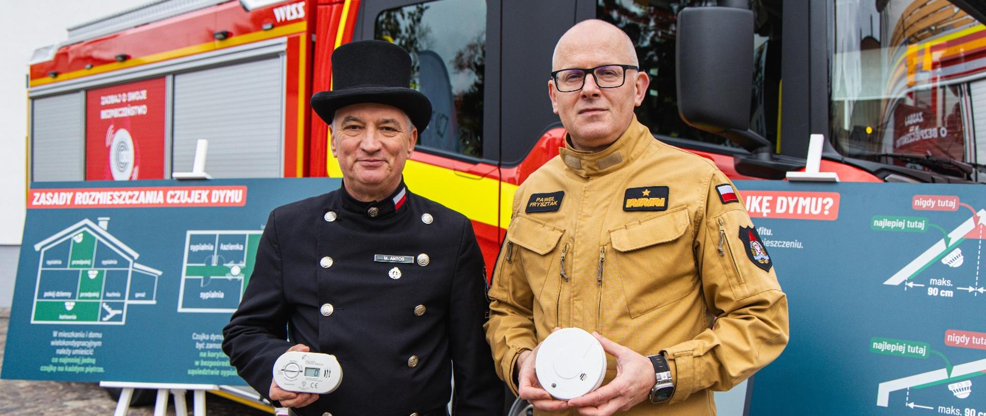 Strażak PSP w żółtym mundurze służbowym oraz kominiarz w czarnym mundurze stoją przed wozem strażackim i prezentują czujki dymu, w tle tablice edukacyjne z zasadami rozmieszczania czujników.