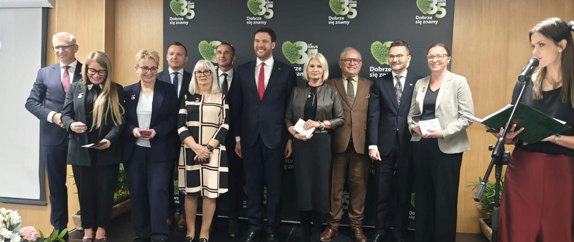 Na tle ścianki z logo 35-lecia KRUS pozuje do zdjęcia 11 osób. Obok przy mikrofonie stoi prowadząca spotkanie rzecznik KRUS. Wszyscy uśmiechają się do zdjęcia.