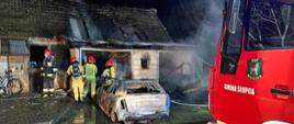 przed budynkiem gospodarczym stoi spalony samochód osobowy przed nim czterech strażaków z boku widoczny przód samochodu strażackiego