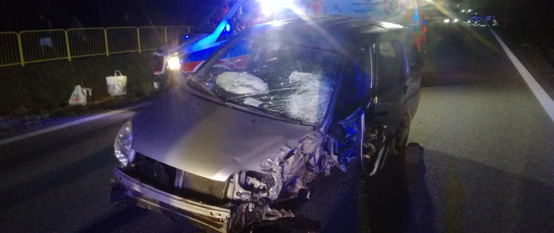 Na zdjęciu zaprezentowany został zniszczony w wyniku zderzenia samochód osobowy marki Subaru, za nim w mroku stoi ambulans. Przy lewym przednim kole widać wyciek płynów ograniczony sorbentem.