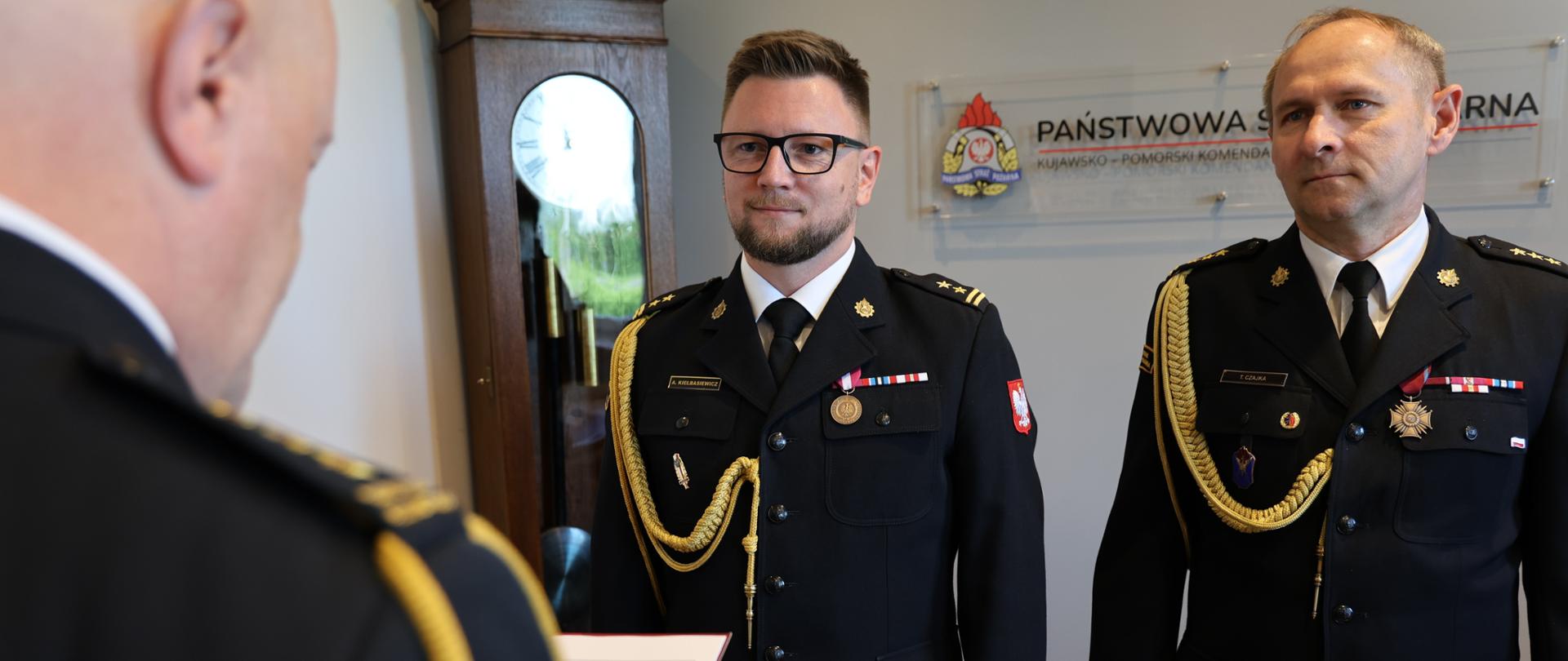 Uroczystość w komendzie straży pożarnej. Dwaj funkcjonariusze w ciemnych mundurach galowych ze sznurami galowymi i baretkami stoją na baczność. W tle widoczne godło oraz napis Państwowa Straż Pożarna Kujawsko-Pomorska Komenda.