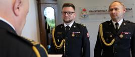 Uroczystość w komendzie straży pożarnej. Dwaj funkcjonariusze w ciemnych mundurach galowych ze sznurami galowymi i baretkami stoją na baczność. W tle widoczne godło oraz napis Państwowa Straż Pożarna Kujawsko-Pomorska Komenda.