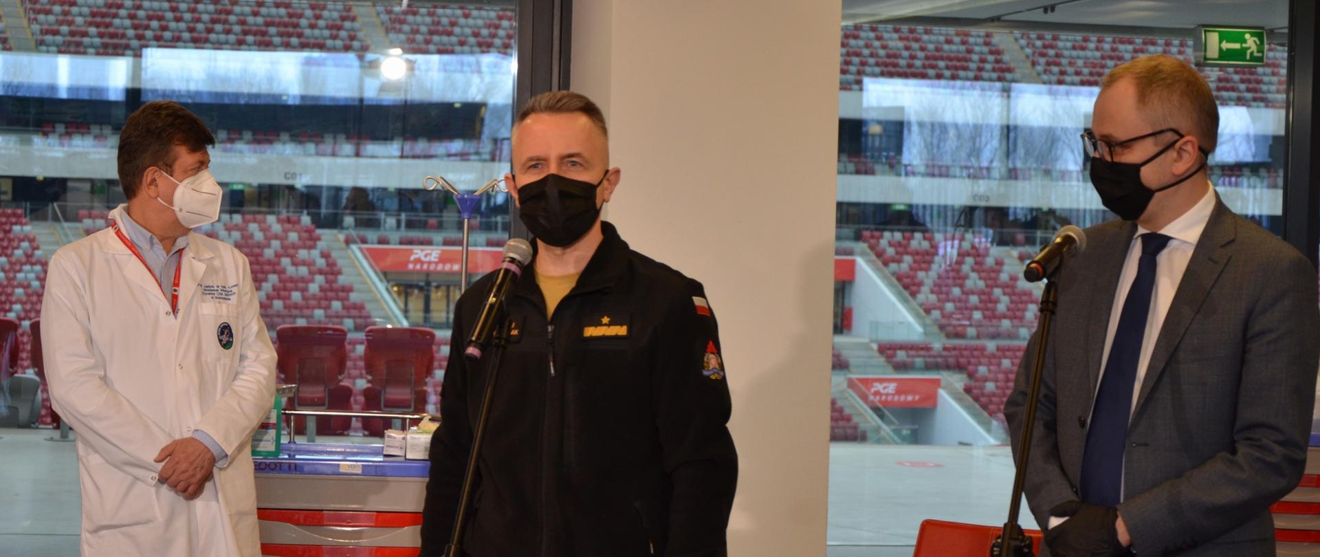 Na pierwszym planie przy mikrofonie stojący Komendant Główny Państwowej Straży Pożarnej nadbrygadier Andrzej Bartkowiak. Przemawia z okazji inauguracji programu szczepień przeciw COVID-19 dla służb mundurowych. Komendant występuje w nowym typie umundurowania (spodnie w kolorze piaskowym, polar w kolorze czarnym). Po prawej stronie zdjęcia, również przy mikrofonie, mężczyzna w szarym garniturze, białej koszuli i granatowym krawacie. Po lewej stronie zdjęcia mężczyna z obsługi medycznej, spogląda w swoją lewą stronę. Jest ubrany w biały kitel. Wszyscy Panowie mają na twarzach maseczki. W tle za oknami widać trybuny Stadionu Narodowego.