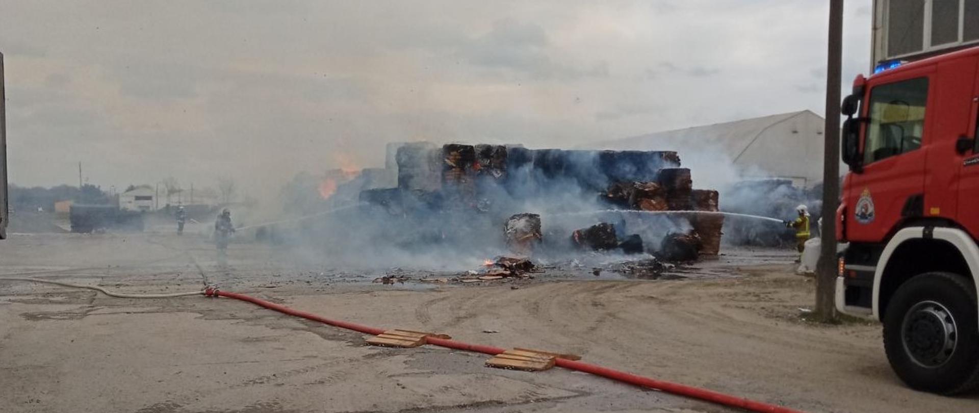 W centralnej części płonąca hałda, wokół której ustawieni są strażacy trzymający węże i gaszący skład makulatury. Po prawej stronie widoczny samochód gaśniczy, a za nim widoczna część budynku. 