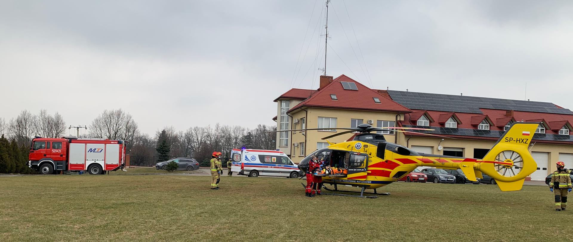 Na zdjęciu widać żółto- czerwony helikopter stojący na trawniku, za nim znajduję się budynek straży pożarnej w Rawie Maz. po lewej stroni znajduję się wóz strażacki oraz samochód karetki pogotowia. Po lewej stronie helikoptera znajdują się dwaj strażacy, a po prawej jeden. Dwóch ratowników medycznych wkłada na noszach poszkodowanego do helikoptera.