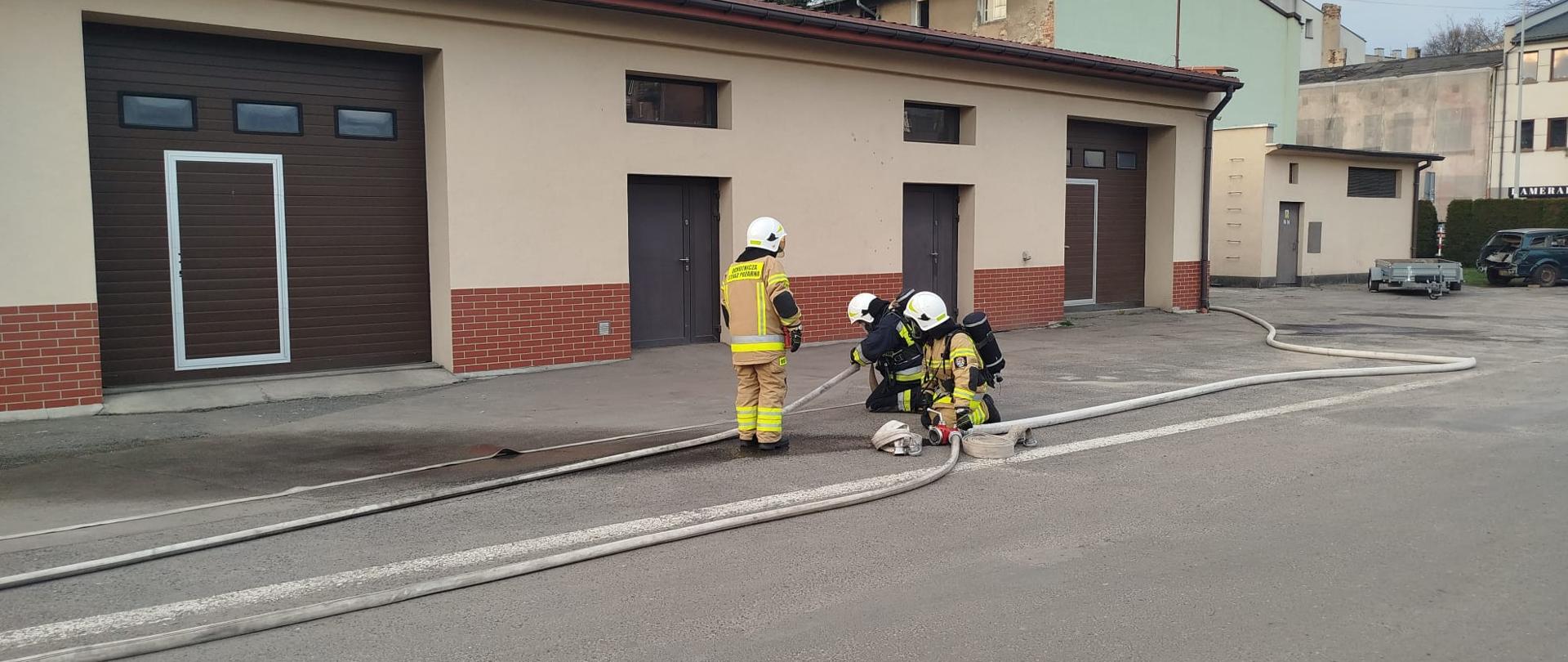 Na zdjęciu widać fragment placu przed budynkiem z kilkoma dużymi bramami garażowymi. Na utwardzonej nawierzchni rozłożone są długie odcinki białych węży strażackich. Dwie osoby w pełnym ubraniu ochronnym strażaka, w jasnożółtych kombinezonach i białych hełmach, pracują przy sprzęcie. Jedna z osób stoi przy wężu, a druga klęczy obok rozdzielacza wodnego. W tle widoczne są budynki, latarnia uliczna oraz zaparkowany samochód.