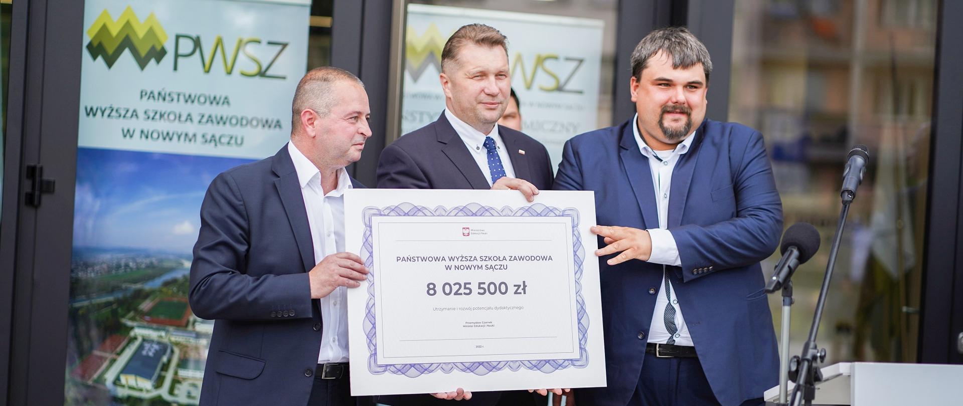 Minister stoi z dwoma ludźmi w garniturach, pokazują wielki symboliczny czek z napisem 8 025 500 zł.