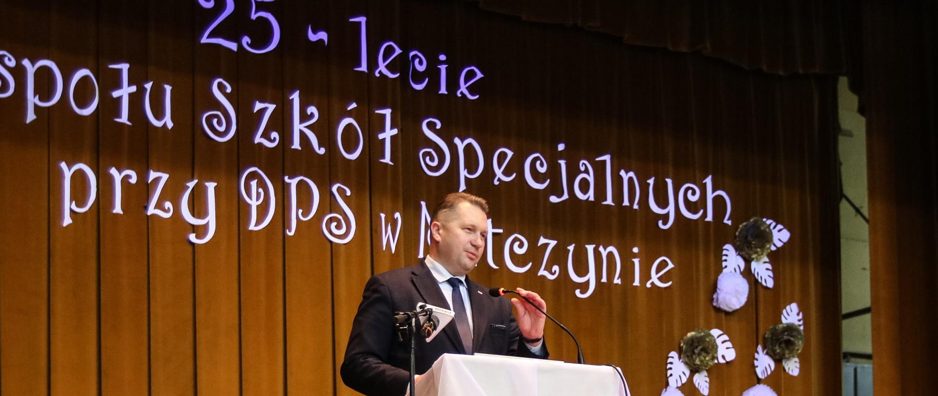 Minister Czarnek stoi przy mównicy i mówi do mikrofonu, za nim złota kotara a na niej niebieski napis 25-lecie Zespołu Szkól specjalnych przy DPS w Matczynie.