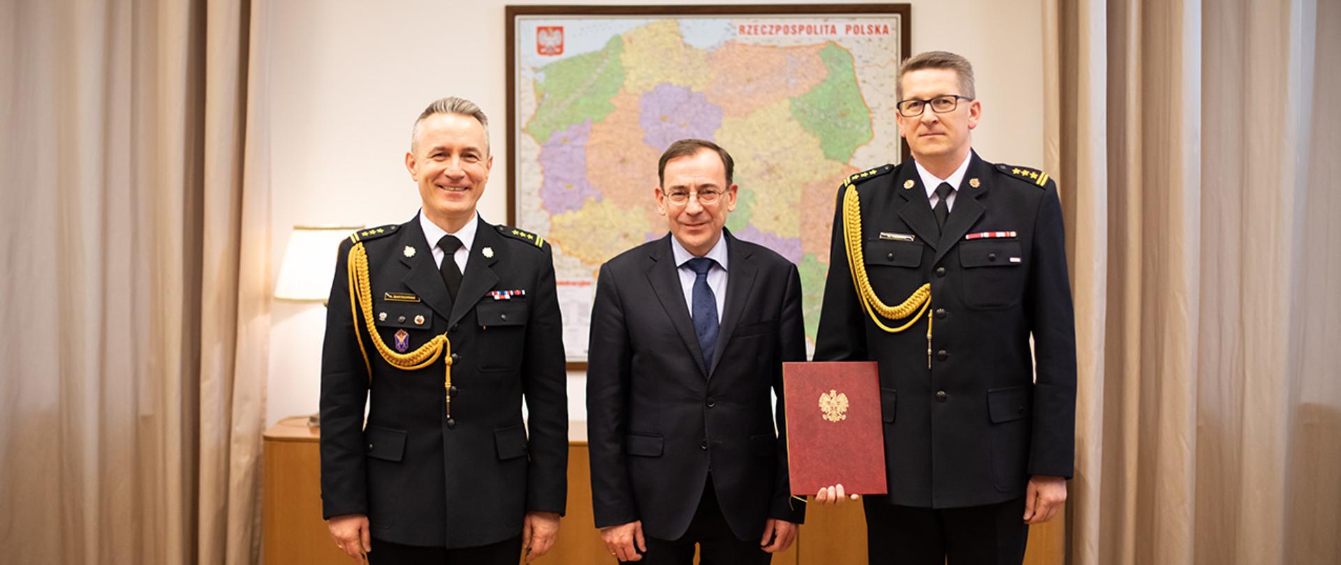 Na zdjęciu widać Mariusza Kamińskiego ministra SWiA, st. bryg. Andrzeja Bartkowiaka komendanta głównego PSP i st. bryg. Krzysztofa Hejduka nowo mianowanego zastępcy komendanta głównego PSP trzymającego sztywną okładkę na dokumenty z aktem mianowania pozujących do wsplnego zdjęcia. W tle widać mapę polityczną Polski.