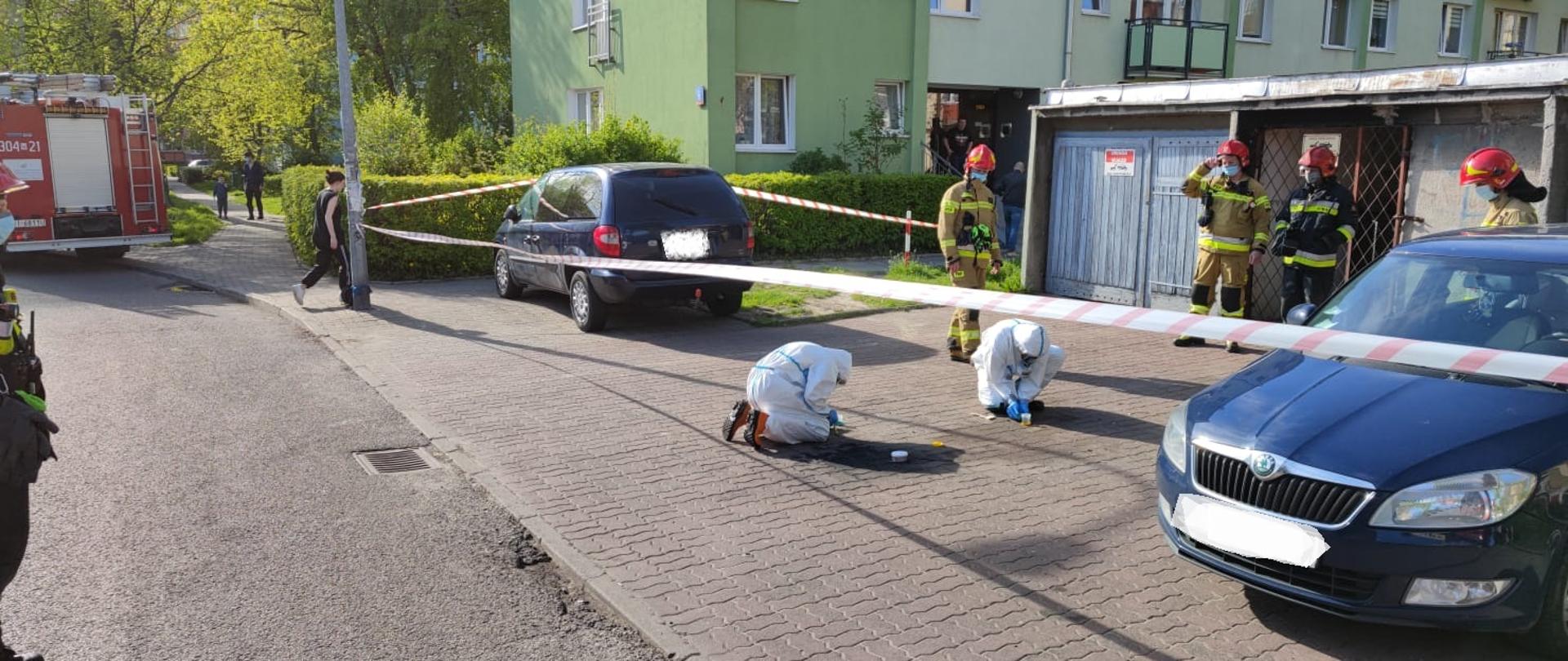 Na zdjęciu widać wygrodzony teren działań biało-czerwoną taśmą ostrzegawczą . Na pierwszym planie dwóch strażaków ubranych w białe ubrania ochronne , żółte buty chemoodporne oraz rękawiczki lateksowe . Strażacy zbierają rozlaną substancję (rtęć) do pojemników. Na zdjęciu widać tez innych strażaków w piaskowych i granatowo żółtych ubraniach którzy zabezpieczają miejsce zdarzenia.